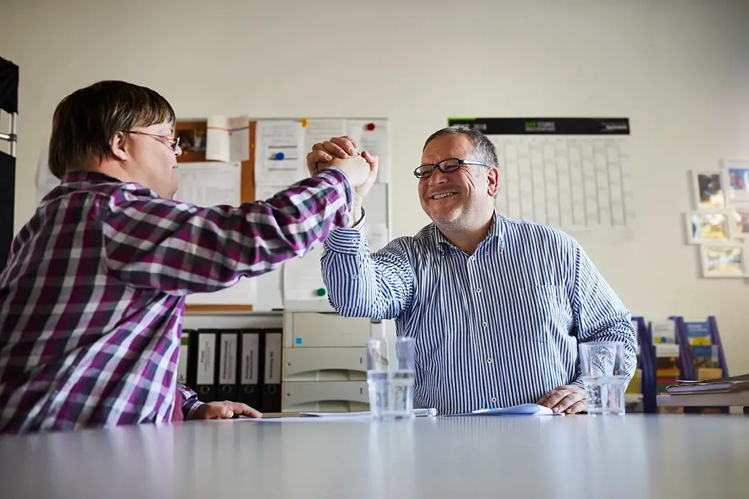Ein junger Mann mit Down-Syndrom und ein Betreuer geben sich mit einem Lächeln ein High-Five über den Tisch – ein starkes Zeichen für Augenhöhe, Vertrauen und Motivation. Genau diese wertschätzende Atmosphäre macht eine Ausbildung oder ein Studium bei der Lebenshilfe Herford so besonders und sinnstiftend.