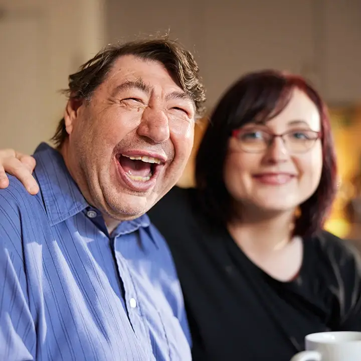 Ein lachender Mann wird herzlich von einer Frau mit Brille umarmt – das Bild zeigt, wie echte Verbindung und Vertrauen die tägliche Arbeit prägen. Genau diese zwischenmenschliche Nähe macht eine Ausbildung oder ein Studium bei der Lebenshilfe Herford zu etwas ganz Besonderem.