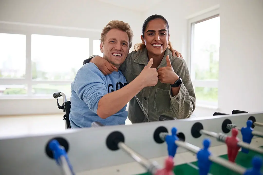 Ein junger Mann im Rollstuhl und eine Frau stehen Arm in Arm vor einem Kickertisch und zeigen beide Daumen hoch – voller Energie, Freude und Zusammenhalt. Für alle, die mit einem Direkteinstieg bei der Lebenshilfe Herford sofort durchstarten und etwas bewegen möchten.