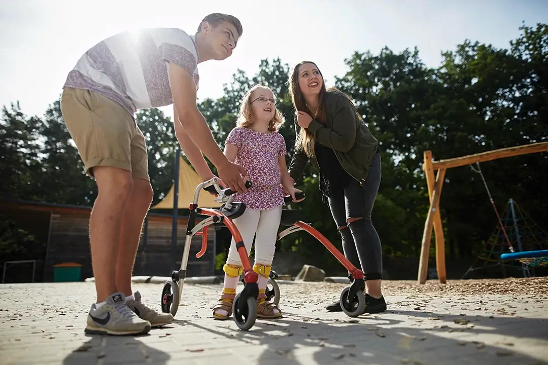 Ein junges Mädchen mit Gehhilfe wird auf einem Spielplatz liebevoll von zwei jungen Erwachsenen begleitet – ein bewegender Moment voller Vertrauen, Unterstützung und Zukunftsfreude. Perfekt für alle, die mit einem Direkteinstieg bei der Lebenshilfe Herford aktiv etwas bewirken wollen.