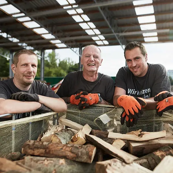 Drei Männer stehen lachend an einer Kiste voller Brennholz in einer Werkhalle – ein authentischer Einblick in die Arbeitswelt und gelebte Teilhabe, wie sie durch ein Praktikum bei der Lebenshilfe Herford möglich wird.