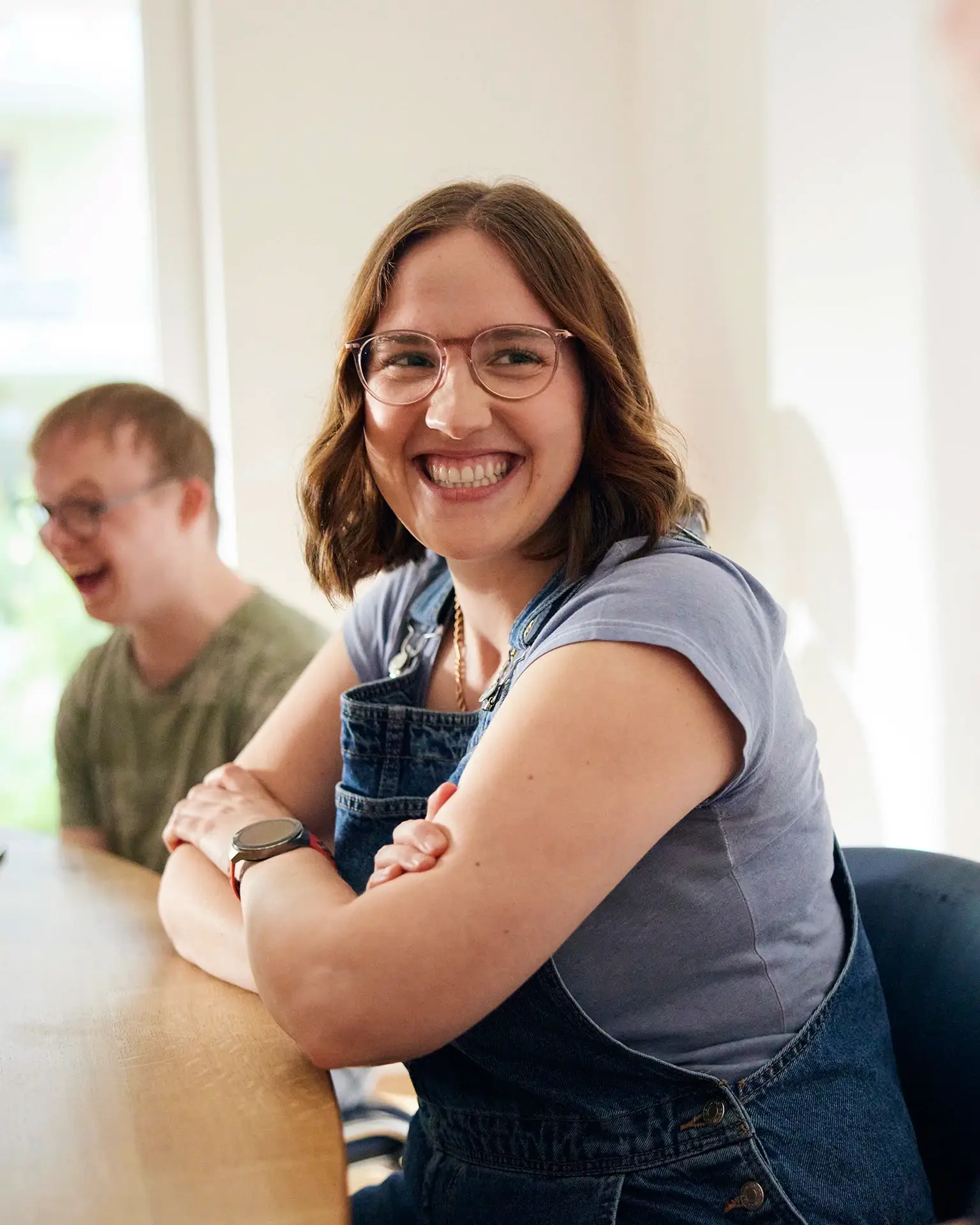 Eine junge Frau mit Brille sitzt strahlend an einem Tisch, im Hintergrund lacht ein junger Mann mit Down-Syndrom &ndash; ein authentischer Moment voller Leichtigkeit und Freude im Arbeitsalltag. Die Stellenangebote der Lebenshilfe Herford bieten nicht nur Sinn, sondern auch sp&uuml;rbare Benefits: ein starkes Team, pers&ouml;nliche Entwicklung und ein Arbeitsplatz mit Herz.