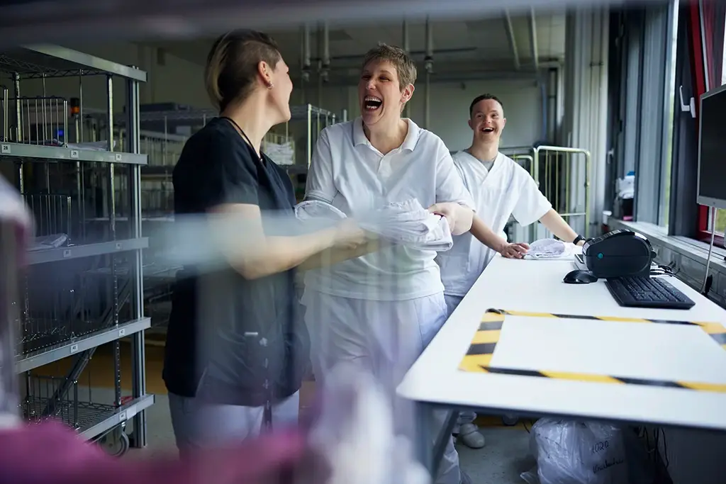 Drei Mitarbeitende in Arbeitskleidung lachen gemeinsam in einer Wäscherei. Die Szene zeigt Teamgeist, Freude an der Arbeit und gegenseitige Unterstützung. Das Bild steht für ein wertschätzendes Miteinander und die gelebte Inklusion innerhalb der Karriere bei der Lebenshilfe Herford.