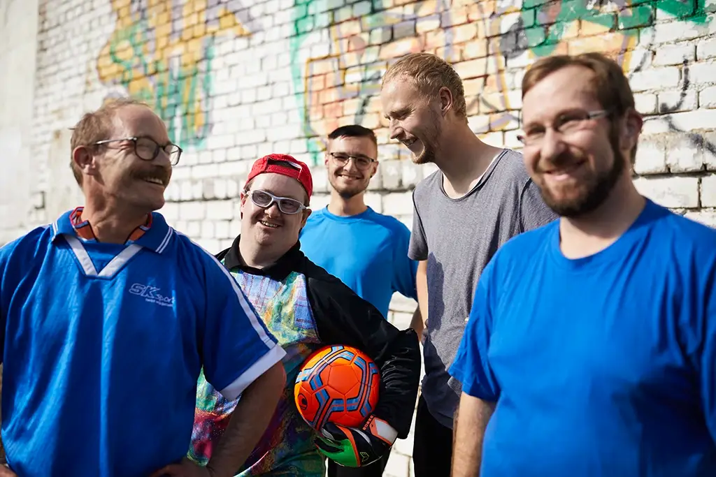 Eine fröhliche Gruppe von fünf Männern steht vor einer bemalten Mauer, einer hält einen Fußball in der Hand. Alle lachen und genießen den Moment gemeinsamer Aktivität. Das Bild symbolisiert Gemeinschaft, Lebensfreude und Inklusion – Werte, die die Karriere bei der Lebenshilfe Herford prägen.