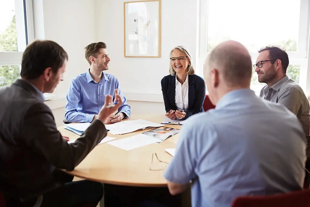 Mehrere Personen sitzen gemeinsam an einem runden Tisch und führen ein positives, lebendiges Gespräch. Alle wirken konzentriert und lachen miteinander. Das Bild steht für Teamarbeit, Vertrauen und eine offene Unternehmenskultur – Werte, die die Karriere bei der Lebenshilfe Herford prägen.