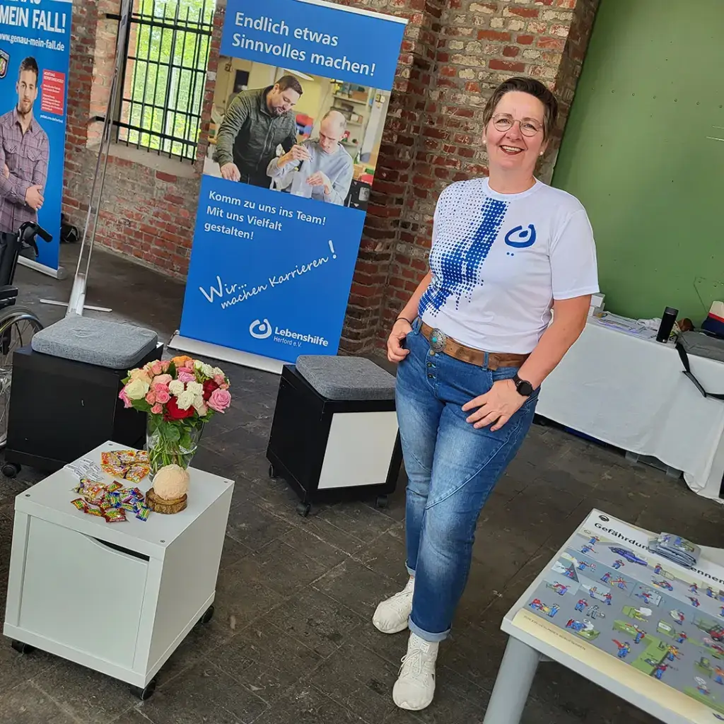 Eine Mitarbeiterin der Lebenshilfe Herford steht lächelnd an einem Infostand mit Flyern, Blumen und Werbematerialien. Im Hintergrund sind Roll-ups mit der Aufschrift „Endlich etwas Sinnvolles machen!“ und „Wir machen Karrieren!“ zu sehen. Das Bild vermittelt Offenheit, Herzlichkeit und die Einladung, Teil eines vielfältigen Teams zu werden.