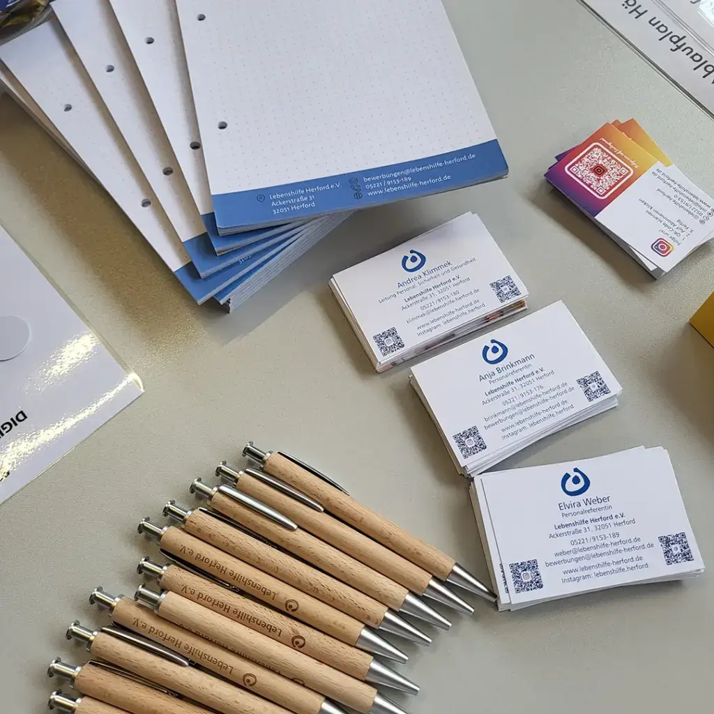 Auf einem Tisch liegen mehrere Schreibblöcke, Holzkugelschreiber mit der Gravur „Lebenshilfe Herford e. V.“ und Visitenkarten von Mitarbeitenden der Lebenshilfe Herford. Die Materialien zeigen das einheitliche, professionelle Erscheinungsbild der Organisation und vermitteln Vertrauen, Nähe und Struktur.