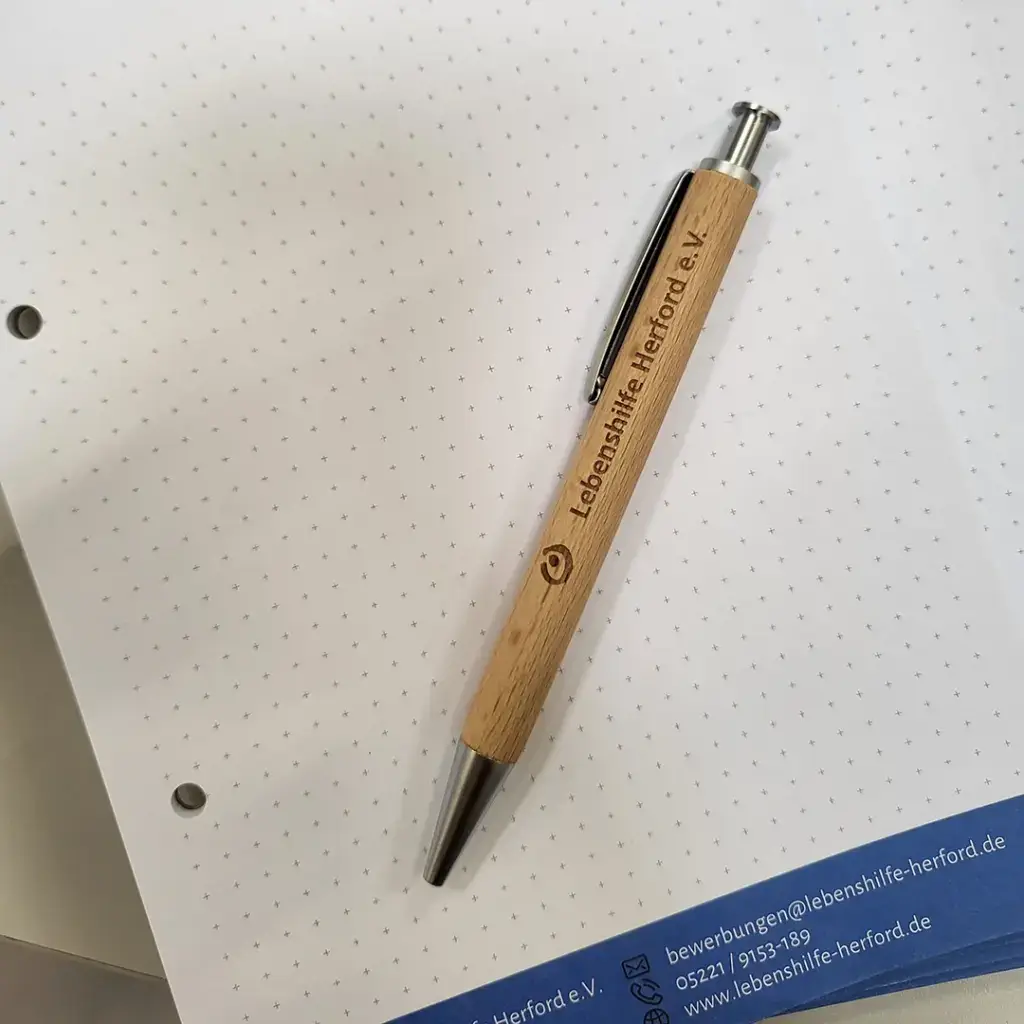 Ein Holz-Kugelschreiber mit der Gravur „Lebenshilfe Herford e. V.“ liegt auf einem karierten Notizblatt. Am unteren Rand ist ein Blatt mit dem Logo und den Kontaktdaten der Lebenshilfe Herford zu sehen. Das Bild vermittelt Professionalität, Nachhaltigkeit und Nähe.