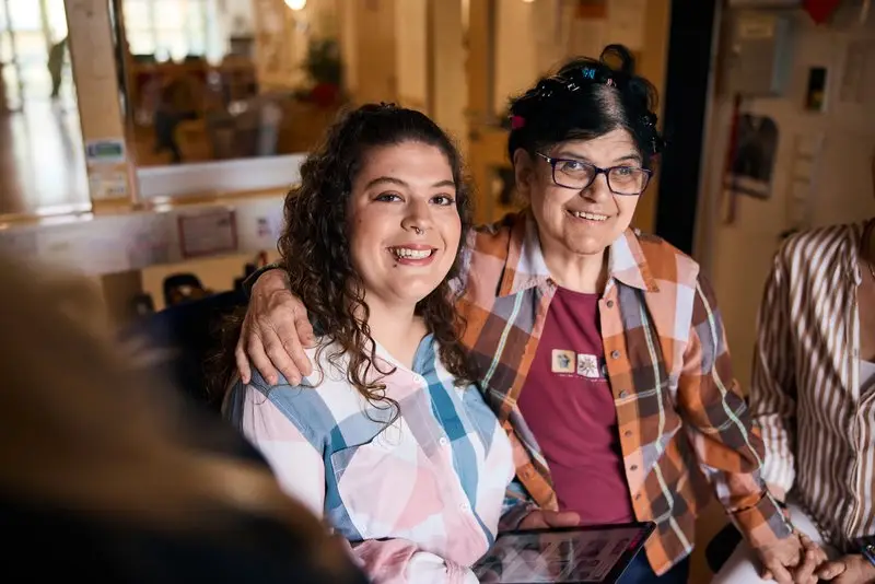 Pflegekraft sitzt lächelnd neben Seniorin und hält ihre Hand in freundlicher Atmosphäre