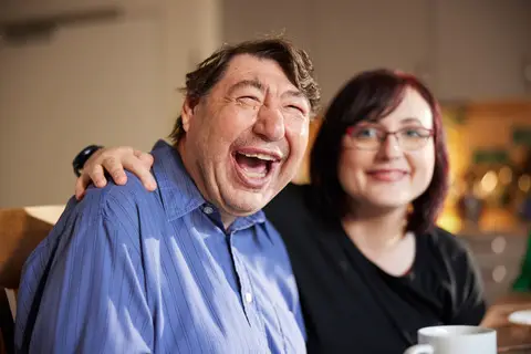 Pflegekraft und Senior lachen gemeinsam bei einer Tasse Kaffee in gemütlicher Umgebung